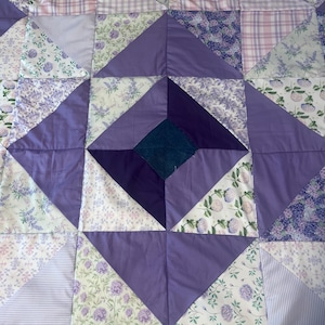 May include: A patchwork quilt featuring a geometric design with a central dark blue square surrounded by triangles and squares in shades of purple, lavender, and white. The fabric patterns include floral prints, stripes, and plaid designs.