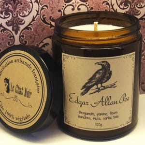 Peut inclure: Une bougie en pot de verre ambré avec un couvercle noir, portant une étiquette avec une illustration de corbeau et le texte Edgar Allan Poe, Bergamote, pomme, fleurs blanches, musc, santal, bois, 320g. Le couvercle indique Fabrication artisanale française, Le Chat Noir, Cire 100% végétale.