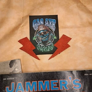 May include: A black and white patch with a skull wearing a blue and white hat and goggles. The skull is holding two wrenches. The text "Gas Axe Chop Shop Supply" is on the patch.