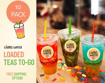 May include: Three clear plastic cups filled with red, green, and blue iced tea, each featuring a Living Water logo sticker, a colored straw, and handwritten labels. The cups are surrounded by scattered colorful candy pieces on a wooden surface.