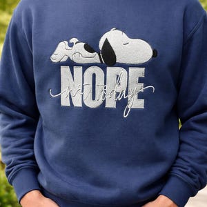 May include: Navy blue sweatshirt featuring Snoopy sleeping with the text "NOPE not today" in white. The sweatshirt has a crew neck and long sleeves. The design is embroidered on the front.