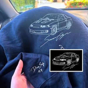 May include: Navy blue sweatshirt featuring a white outline of a car and the text "Soarer Style". The sleeve displays "Drive Safe" and "AY" in white script. A smaller image of the car is in the corner.