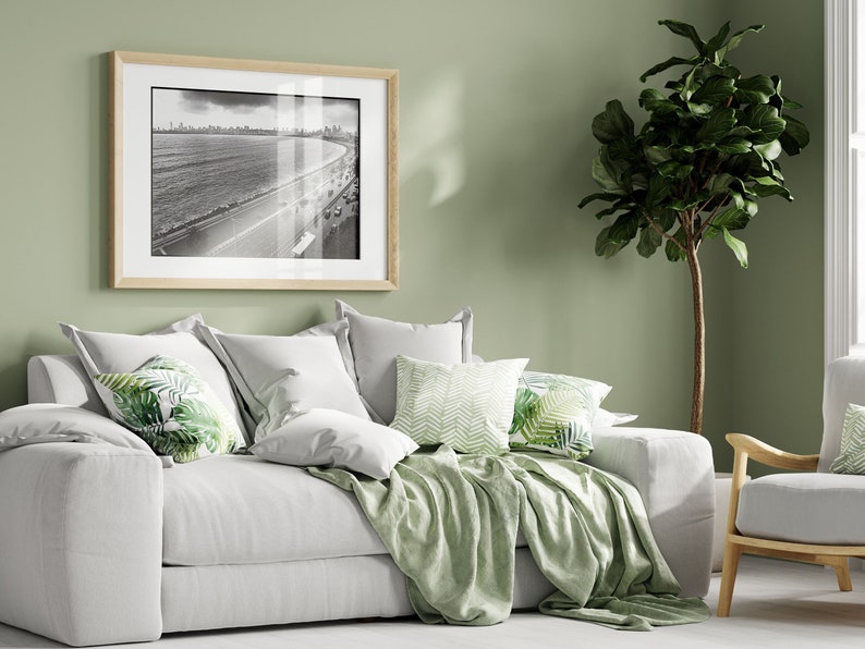 Może przedstawiać: Biała sofa z zielonym kocem i poduszkami z tropikalnymi wzorami liści. Za sofą na ścianie wisi oprawiona czarno-biała fotografia przedstawiająca scenę plażową. W rogu pokoju stoi zielona roślina w doniczce.