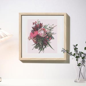 Könnte beinhalten: Gerahmtes Kreuzstich-Kunstwerk mit einem Bouquet aus rosa und roten Blumen mit grünem Laub. Das Kunstwerk befindet sich in einem hellen Holzrahmen und hängt an einer weißen Wand. Eine Lampe und eine Vase mit Blumen sind ebenfalls zu sehen.