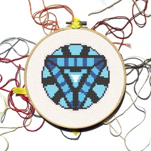 May include: A cross-stitch embroidery hoop featuring a geometric design in shades of blue, black, and white. The design is centered on a white fabric, secured within a wooden hoop. Various colored threads surround the hoop.