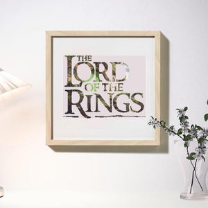 May include: A framed print featuring the text "THE LORD OF THE RINGS" in a stylized font with a nature-inspired design. The print is in a light wood frame and hangs on a white wall. A lamp and a vase of flowers are also visible.