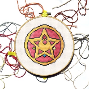 May include: A cross-stitch embroidery hoop with a star design in yellow, pink, and black. The design is on cream fabric, held in a wooden hoop. Various coloured threads, including orange and grey, are scattered around the hoop.
