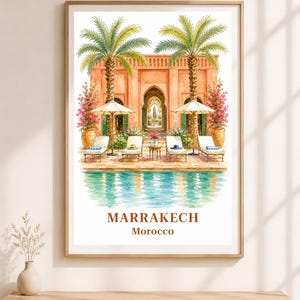 May include: A framed watercolor print depicting a swimming pool in front of a terracotta-colored building with palm trees, lounge chairs, and umbrellas. The text MARRAKECH Morocco is printed in brown at the bottom of the white-bordered artwork.