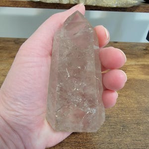 Op de afbeelding: Een heldere kwartskristaltoren met een lichtbruine tint. Het kristal wordt in een hand gehouden, waardoor het gepolijste oppervlak en de natuurlijke insluitsels zichtbaar worden. De torenvorm loopt taps toe naar een punt, wat de geometrische vorm benadrukt.