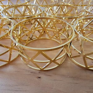 Puede incluir: Una colección de portavelas de metal dorado. Cada soporte tiene una forma cilíndrica con un diseño geométrico abierto. Los soportes están dispuestos sobre una superficie marrón claro, creando una exhibición visualmente atractiva. El diseño presenta triángulos interconectados.