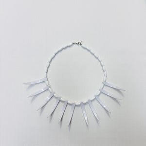 May include: A white necklace with a series of white, elongated, oval beads forming a circular shape. Attached to the necklace are several white, pointed, blade-like pendants, evenly spaced around the lower half of the necklace.