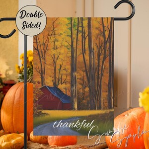 May include: A double-sided garden flag with a red barn in a forest with fall foliage. The text "thankful" is written on the bottom of the flag.