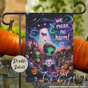 May include: A rectangular garden flag featuring a colorful illustration of green aliens, a UFO abducting a cow, and pumpkins. The text reads We mean no harm! A circular beige tag attached to the black metal stand displays the text Double Sided!