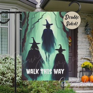 May include: Double-sided Halloween garden flag featuring three witch silhouettes walking through a misty forest. The flag has the text "WALK THIS WAY" in white. The flag is displayed on a black metal stand in front of a house.
