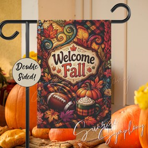 May include: A rectangular garden flag hanging on a black metal stand, featuring a colorful autumn design with the text Welcome Fall. The illustration includes a football, a pumpkin, a mug of hot cocoa, and fall leaves. A circular label reads Double Sided!