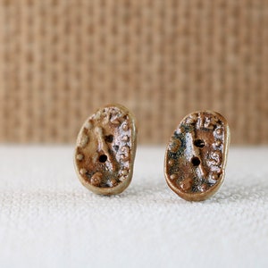 May include: A pair of ceramic earrings shaped like clock faces with brown and white glaze. The clock faces have Roman numeral markings and a small hole in the center.