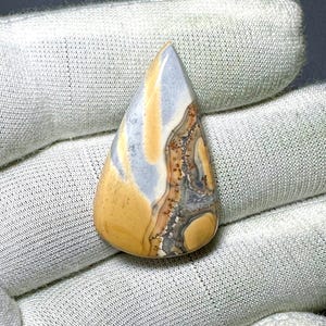 Puede incluir: Una gema pulida en forma de lágrima. La piedra presenta una mezcla de colores, incluyendo azul claro, beige y marrón, con patrones intrincados. La gema se utiliza probablemente para joyería o fines decorativos.