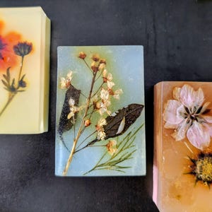 May include: Three rectangular bars of soap with dried flowers encased in clear resin. One bar features an orange flower, another has white flowers and a dark leaf, and the third has a pink flower and a yellow center.