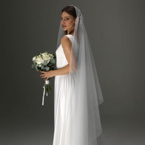 May include: A bride in a white sleeveless wedding dress and veil, holding a bouquet of white roses and greenery. The veil is sheer and falls to the floor. The dress has a simple, elegant design.