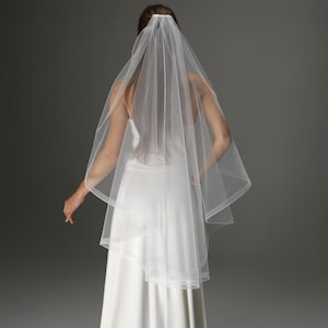 May include: A bride in a white wedding dress and a sheer, two-tiered veil. The veil is secured at the crown and falls over the shoulders. The floor-length dress has a simple, elegant design, suitable for a wedding.