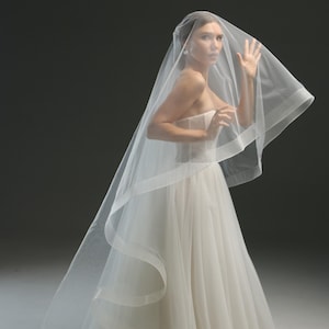 May include: A strapless white wedding gown with a flowing skirt and a sheer veil. The veil drapes over the shoulders and head, creating a soft, ethereal look. The dress has a fitted bodice and a full skirt.