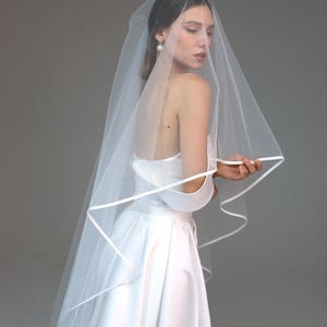 May include: A bride in a white satin wedding dress and a sheer veil with a satin trim. The veil drapes over the shoulders and back. The bride is wearing a pearl earring.