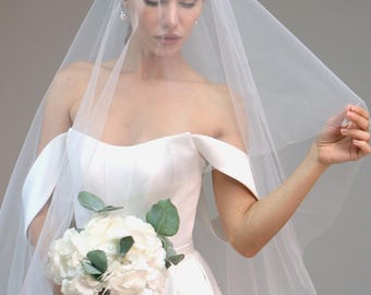 May include: A bride wearing a white off-the-shoulder wedding dress with a long veil and holding a bouquet of white flowers.