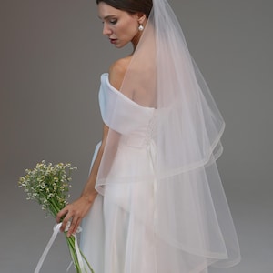 Pode incluir: Uma noiva usando um vestido de noiva branco de ombro caído com um véu longo e fluido. Ela está segurando um buquê de flores brancas.