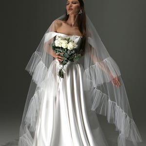 Può includere: Abito da sposa in raso bianco con corpetto senza spalline e gonna fluida. La sposa tiene in mano un bouquet di rose bianche e indossa un lungo velo trasparente con finiture a balze. L'abito è lungo.