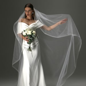May include: A white satin wedding dress with an off-the-shoulder neckline and a flowing skirt. The bride holds a bouquet of white roses and greenery. A sheer veil drapes over the head and extends to the side.