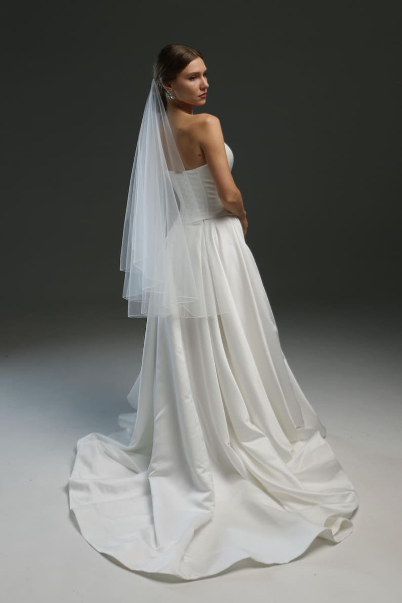 K&ouml;nnte beinhalten: Eine Braut in einem wei&szlig;en, tr&auml;gerlosen Brautkleid mit langem Schleier und Schleppe. Das Kleid hat ein figurbetontes Oberteil und einen flie&szlig;enden Rock.