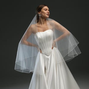 K&ouml;nnte beinhalten: Ein wei&szlig;es, tr&auml;gerloses Brautkleid mit einem figurbetonten Oberteil und einem flie&szlig;enden Rock. Das Kleid wird mit einem wei&szlig;en T&uuml;llschleier kombiniert.