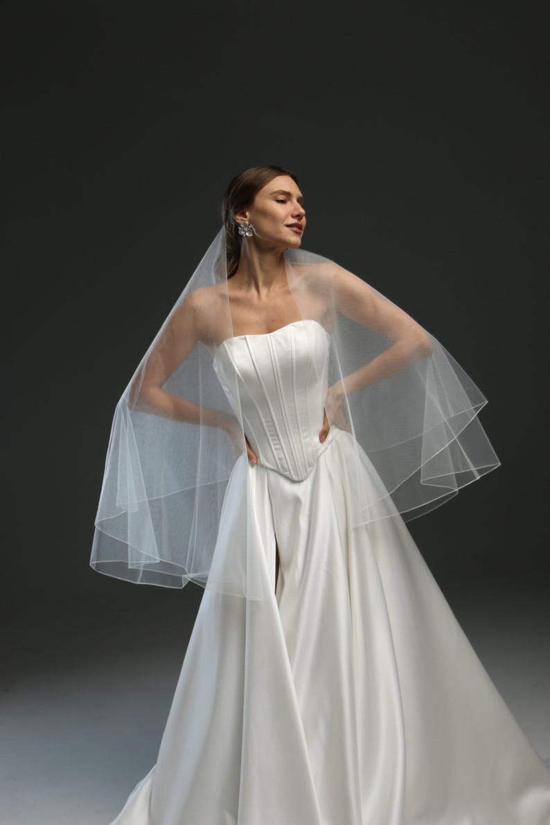 K&ouml;nnte beinhalten: Ein wei&szlig;es, tr&auml;gerloses Brautkleid mit einem figurbetonten Oberteil und einem flie&szlig;enden Rock. Das Kleid wird mit einem wei&szlig;en T&uuml;llschleier kombiniert.