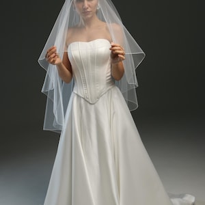 K&ouml;nnte beinhalten: Eine Braut in einem wei&szlig;en, tr&auml;gerlosen Brautkleid mit langem Schleier. Das Kleid hat ein tailliertes Oberteil und einen flie&szlig;enden Rock.