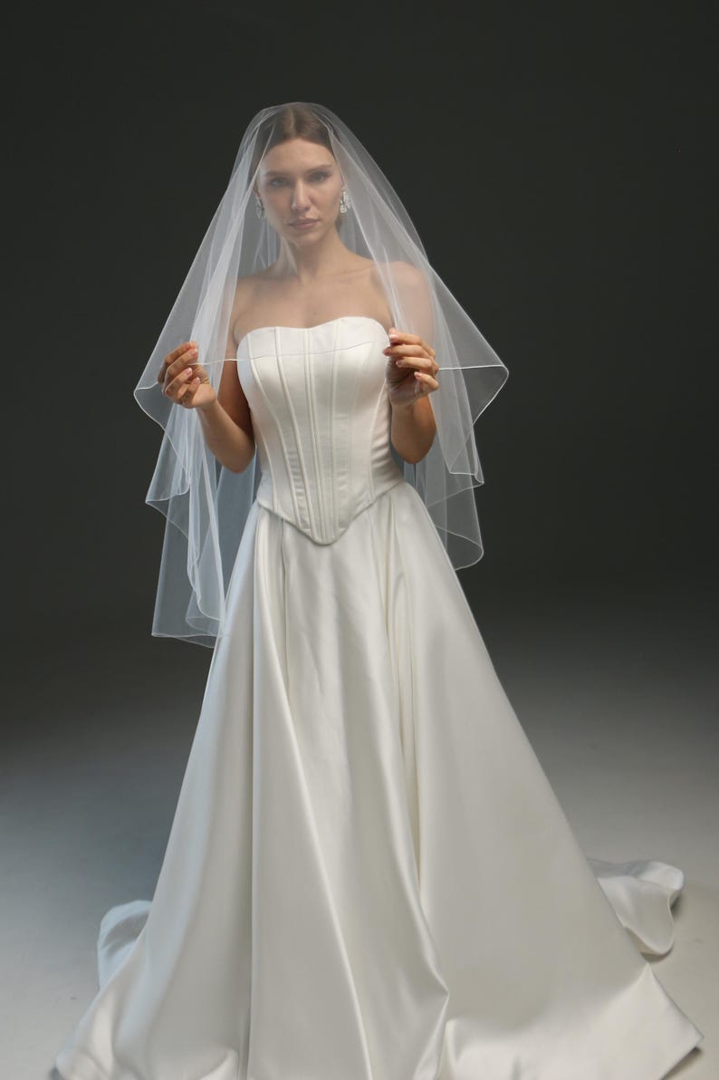 K&ouml;nnte beinhalten: Eine Braut in einem wei&szlig;en, tr&auml;gerlosen Brautkleid mit langem Schleier. Das Kleid hat ein tailliertes Oberteil und einen flie&szlig;enden Rock.