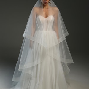May include: A white wedding dress with a strapless bodice and layered skirt, paired with a sheer veil. The veil cascades over the shoulders and back. The dress has a fitted bodice and a flowing skirt, creating an elegant bridal silhouette.