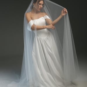 Puede incluir: Un vestido de novia blanco con hombros descubiertos, falda fluida y velo transparente. El vestido presenta un corpiño ajustado y un diseño suave y elegante. El velo está drapeado sobre los hombros, creando un look romántico y etéreo.