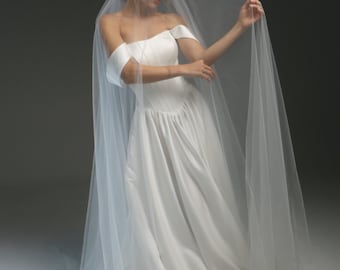 Op de afbeelding: Een witte trouwjurk met off-the-shoulder design en een zwierige rok, gecombineerd met een doorschijnende sluier. De jurk heeft een getailleerd lijfje en een zachte, elegante stijl. De sluier is over de schouders gedrapeerd, wat een romantische uitstraling creëert.
