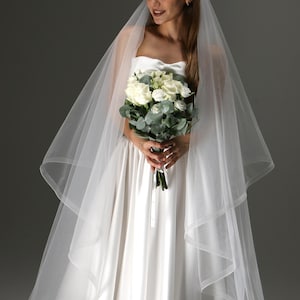 May include: A bride in a white strapless wedding dress and veil holds a bouquet of white roses and greenery. The veil cascades down the shoulders, creating a soft, ethereal look. The dress has a simple, elegant design.
