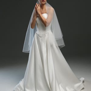 K&ouml;nnte beinhalten: Eine Braut in einem wei&szlig;en, tr&auml;gerlosen Brautkleid mit langem Schleier. Das Kleid hat ein tailliertes Oberteil und einen flie&szlig;enden Rock.