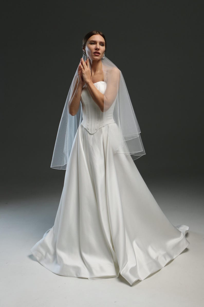 K&ouml;nnte beinhalten: Eine Braut in einem wei&szlig;en, tr&auml;gerlosen Brautkleid mit langem Schleier. Das Kleid hat ein tailliertes Oberteil und einen flie&szlig;enden Rock.