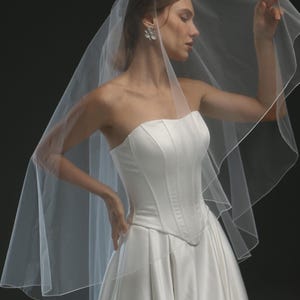 K&ouml;nnte beinhalten: Ein einlagiger Braut-Schleier aus T&uuml;ll mit einem gewellten Saum. Der Schleier ist &uuml;ber den Kopf und die Schultern der Braut gelegt, die ein wei&szlig;es, tr&auml;gerloses Brautkleid tr&auml;gt.