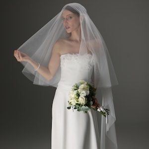Op de afbeelding: Een bruid in een witte strapless trouwjurk houdt een boeket witte rozen en groen vast. Ze draagt een doorschijnende, gelaagde sluier. De jurk heeft een eenvoudig, elegant ontwerp.