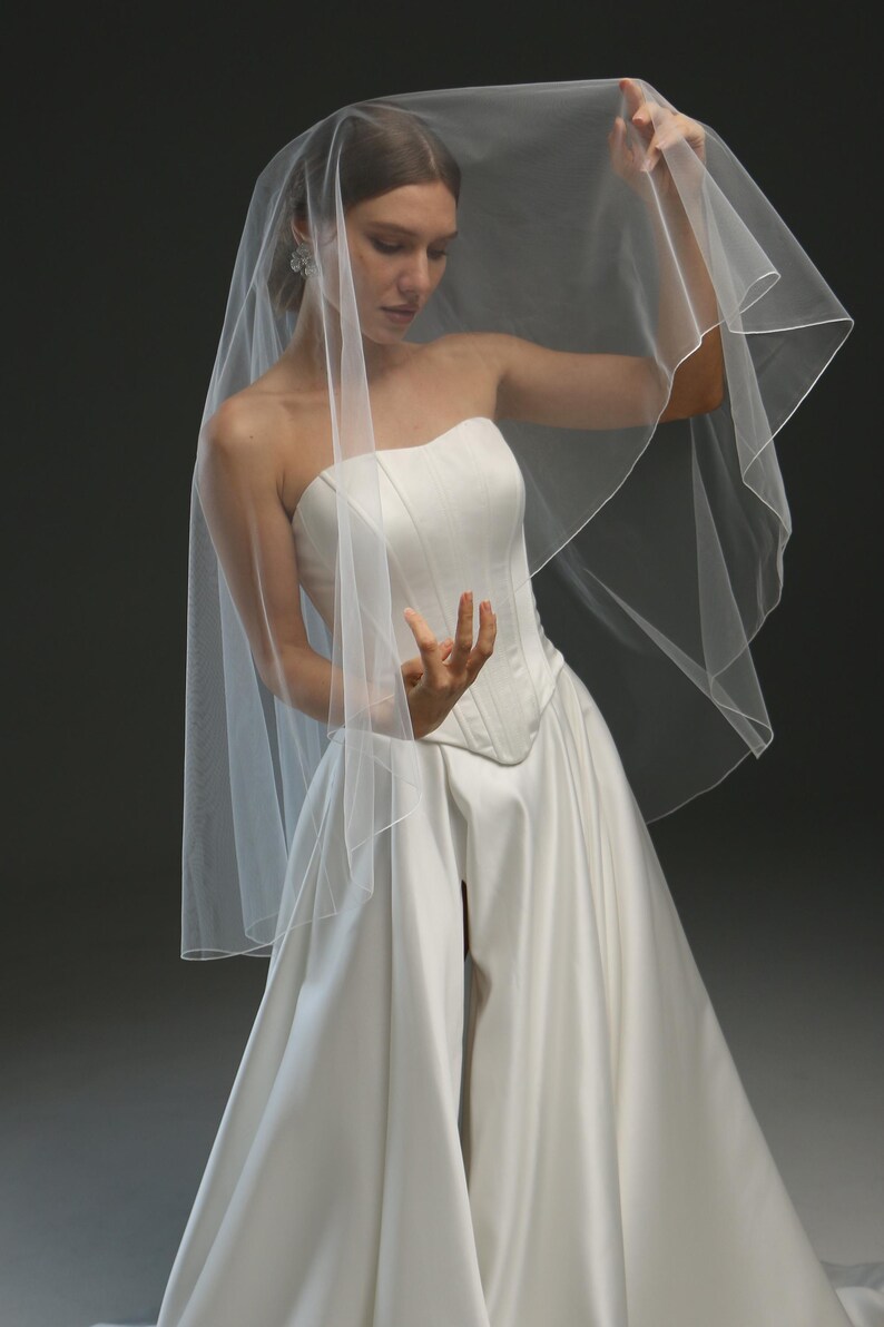 K&ouml;nnte beinhalten: Ein einlagiger Schleier aus T&uuml;ll, der &uuml;ber einem wei&szlig;en tr&auml;gerlosen Brautkleid getragen wird.