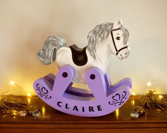 May include: A purple rocking horse with a white horse head and a black saddle. The rocking horse is on a wooden shelf with a name plaque that reads "CLAIRE".