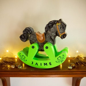 May include: A wooden rocking horse toy with a gray body, brown saddle, and green base. The name "JAIME" is painted on the base. The toy is displayed on a wooden shelf with decorative lights and glass beads.