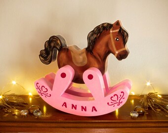 May include: A pink wooden rocking horse with a brown horse head and mane. The rocking horse has a saddle and is decorated with a heart design. The rocking horse is personalized with the name "ANNA".