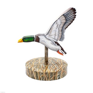 Op de afbeelding: Een houten eendenlokvogel met beschilderde details, in vlucht op een basis met een bruin en beige camouflagepatroon.