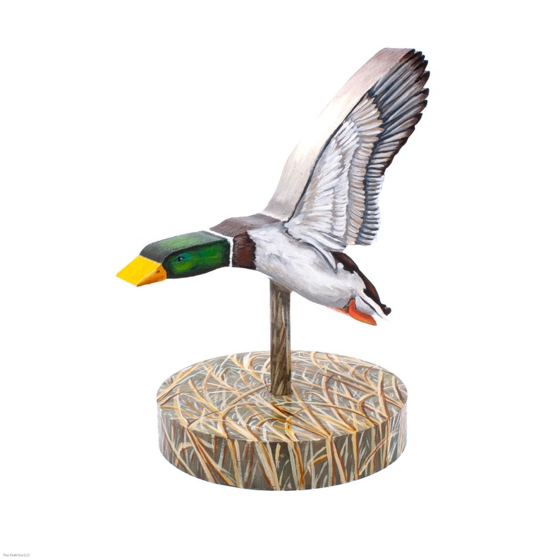Op de afbeelding: Een houten eendenlokvogel met beschilderde details, geperst op een basis met een bruin en beige grasachtig ontwerp.