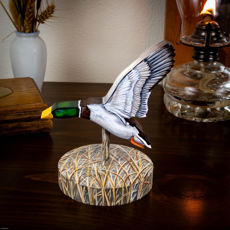 Op de afbeelding: Handgeschilderde houten eendenlokvogel met een groene kop, gele snavel en witte en grijze vleugels. De lokvogel is gemonteerd op een houten voet met een geschilderde afbeelding van riet.
